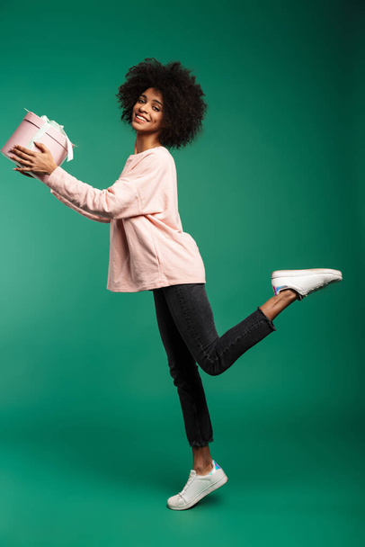 Full length portrait of a smiling young african woman wearing sweatshirt jumping isolated over green background, holding big present box - 写真・画像