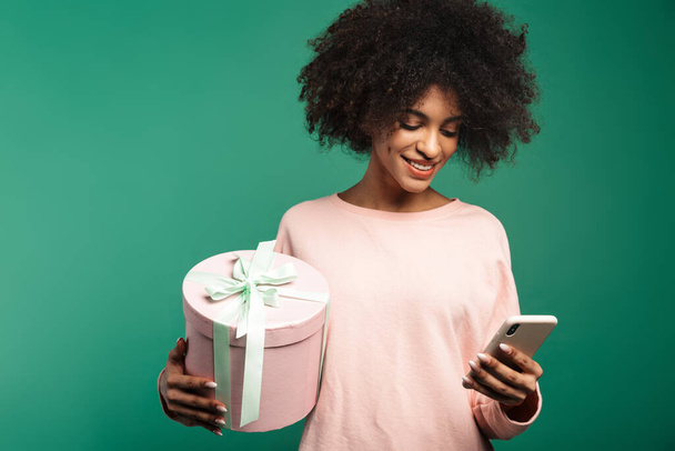 Portrait of a smiling young african woman wearing sweatshirt standing isolated over green background, using mobile phone while holding big present box - Photo, Image