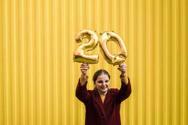 Young beautiful sad girl in burgundy coat and mustard sweater holds in her hands the helium balloons of number 20 against the background of bright yellow wall. Birthday concept in 20 years. - Фото, изображение