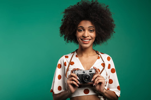 Portrait of a happy young african woman standing isolated over green background, wearing summer clothes, holding photo camera - Foto, Imagem