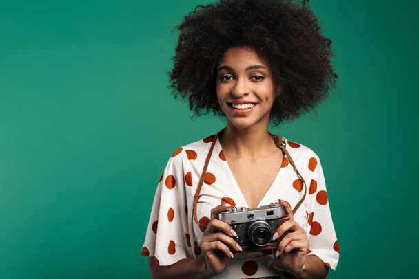 Portrait of a happy young african woman standing isolated over green background, wearing summer clothes, holding photo camera - 写真・画像