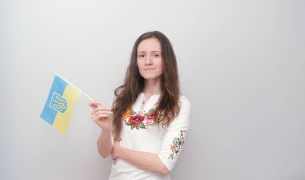 Young beautiful girl in embroidered shirt with Ukrainian flag. Attractive Ukrainian woman wearing traditional Ukrainian embroidery on the grey background.  - Photo, Image