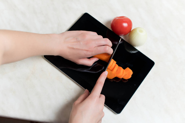 An old tablet serves as a cutting board for cutting vegetables, a woman cuts vegetables on an old tablet computer. Technology obsolescence and new applications - Foto, Imagen