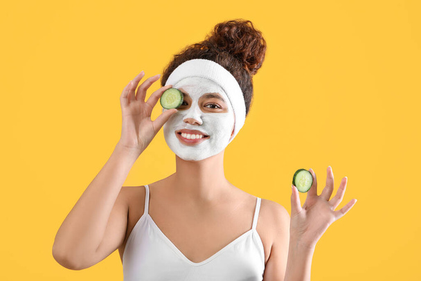 Beautiful young African-American woman with cucumber slices and facial mask on color background - Photo, image