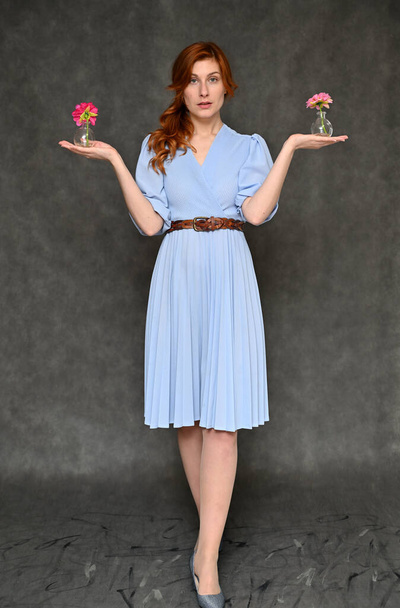 The model demonstrates standing in full growth different poses with a flower in her hands. Vertical portrait of a young pretty red-haired woman in a blue dress on a gray background. - Foto, Bild
