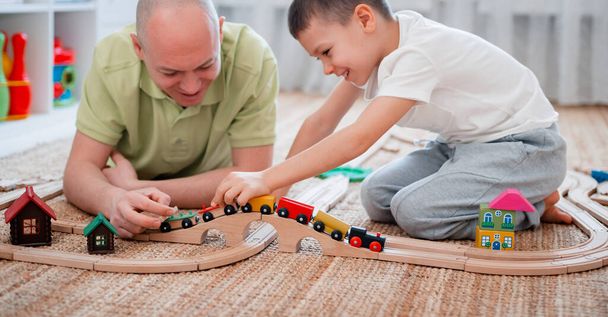 Aile babası ve oğlu oyun odasında oyuncak ahşap bir demiryolu oynuyorlar. Ebeveyn ve çocuğun birlikte zaman geçirmesi kavramı. seçici odak - Fotoğraf, Görsel