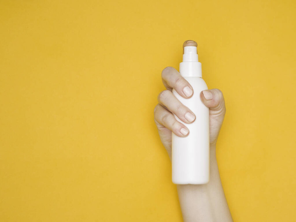 白いプラスチックスプレーボトルを持つ女性の手。ケア化粧品防腐剤。黄色の背景. - 写真・画像