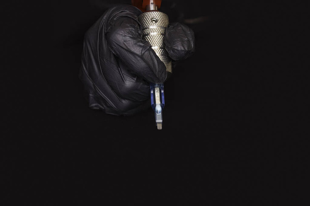 Parte de una máquina rotativa de tatuaje con una aguja en la mano en un guante negro sobre un fondo negro. Vista lateral
 - Foto, Imagen