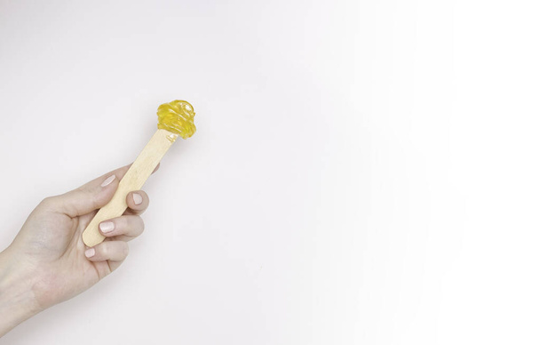 Uma mão de mulher com uma espátula de madeira e pasta de açúcar para depilação. Fundo de luz, espaço livre para texto
. - Foto, Imagem