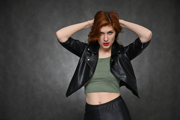 Portrait of a young pretty woman with beautiful hair and excellent make-up in a green T-shirt and a black jacket on a gray background. The model shows different emotions by changing poses. - Foto, afbeelding