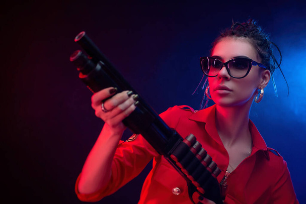 bright brunette in a red jumpsuit with a shotgun on a dark background in neon light - Photo, Image