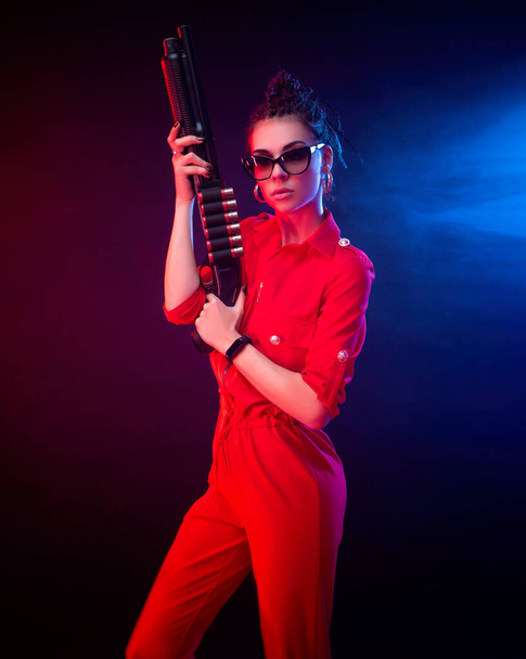 bright brunette in a red jumpsuit with a shotgun on a dark background in neon light - Φωτογραφία, εικόνα