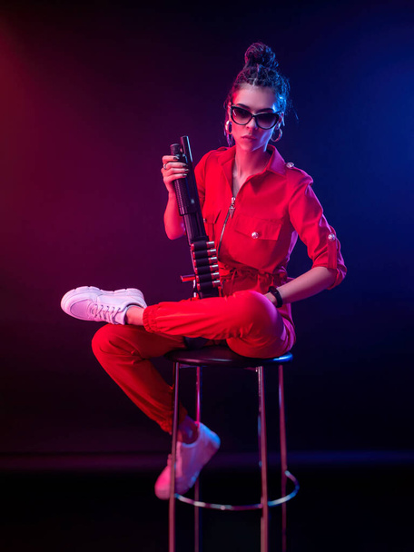 bright brunette in a red jumpsuit with a shotgun on a dark background in neon light - Фото, изображение