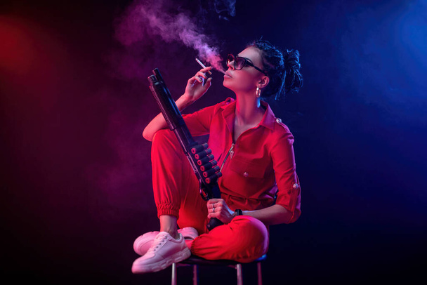 bright brunette in a red jumpsuit with a shotgun on a dark background in neon light - Фото, зображення