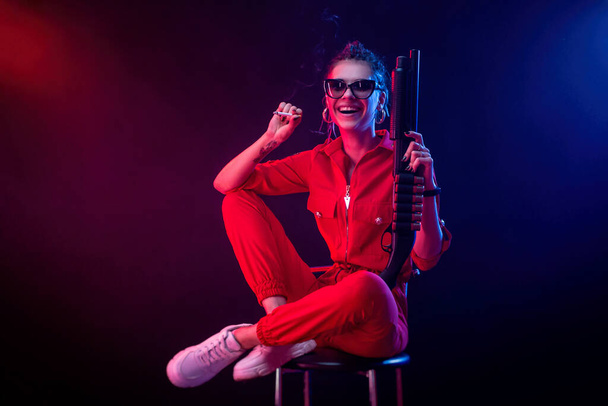 helder brunette in een rood jumpsuit met een shotgun op een donkere achtergrond in neon licht - Foto, afbeelding
