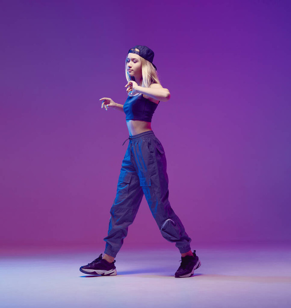 Cute teen girl dancing hip-hop in stylish clothes, a baseball cap, in a Studio with neon lighting. Dance color poster. - Фото, изображение