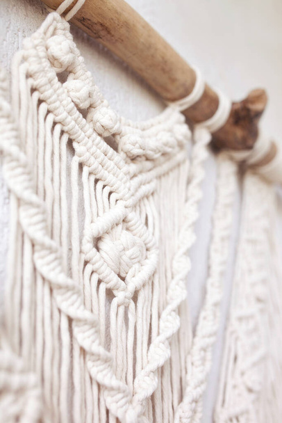 Handmade macrame  100% cotton wall decoration with wooden stick hanging on a white wall.  Macrame braiding and cotton threads. Details close up. Female hobby.   - Foto, imagen