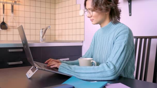 Video of young girl looks screen monitor, typing text on keyboard, writing intently, working, sitting in quarantine during pandemic. Beautiful brunette Bob hair wears blue sweater. coffee mug Notepad - Metraje, vídeo