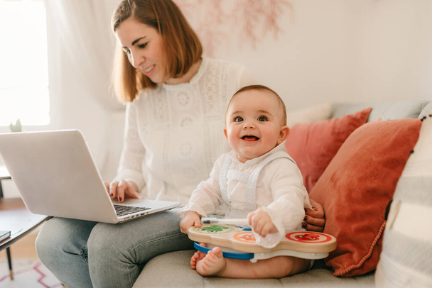 jovem mãe de negócios trabalha em casa com seu laptop, com seu bebê ao seu lado se divertindo
 - Foto, Imagem