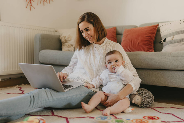 junge Geschäftsfrau arbeitet von zu Hause aus mit ihrem Laptop, mit ihrem Baby an ihrer Seite, das Spaß hat - Foto, Bild