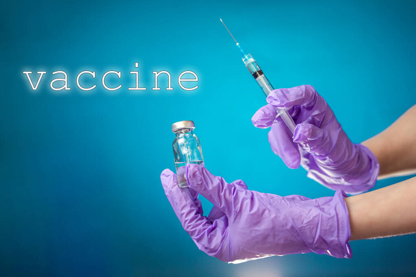 Impfen. Die Hand des Arztes in medizinischen Handschuhen hält ein Fläschchen und eine Spritze für die Injektion. Blauer Hintergrund. Das Konzept von Coronavirus, Impfstoffen und Gesundheit. - Foto, Bild