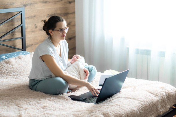 remote work, training, work at home, femininity, technology, leisure, communication online concept - Young cute girl with newborn baby sits on bed with laptop and works remotely, copy space. - Φωτογραφία, εικόνα