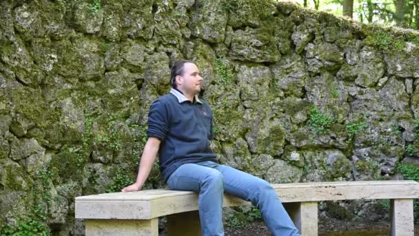 Muž sedí na kamenné lavičce a dívá se, používá své chytré hodinky, zatímco v pozadí je stará zeď jako jednotlivé velké kameny - Záběry, video