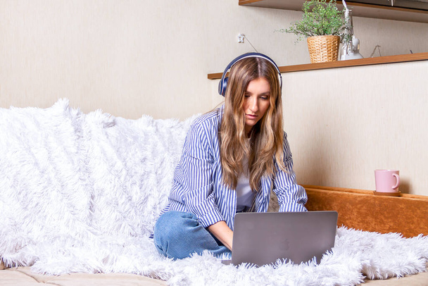 Çevrimiçi eğitim ve çalışma mesafesi. Kulaklıklı, dalgalı saçlı bir iş kadını kanepede çalışıyor ve kahve içiyor. Bilgisayar kullanarak ve müzik dinleyerek. Evde kal. - Fotoğraf, Görsel