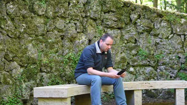 um homem senta-se em um banco de pedra e joga com seu telefone celular e recebe uma chamada enquanto há uma parede velha no fundo como pedras grandes individuais
 - Filmagem, Vídeo