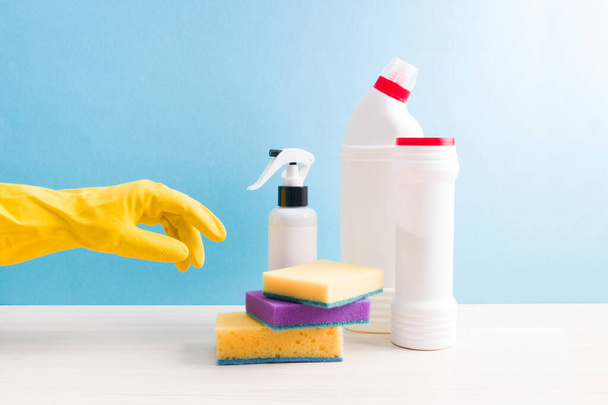 hand in een gele rubberen handschoen reikt naar een reinigingsspons, witte flessen zonder labels met wasmiddelen en spray op tafel, blauwe achtergrond, kopieerruimte - Foto, afbeelding