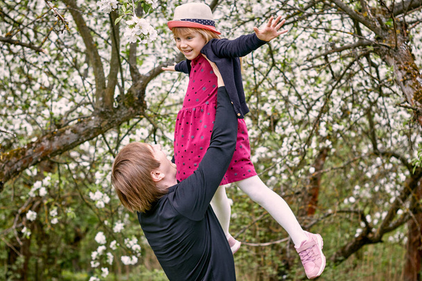 A young father plays with his daughter in a flowering garden. Raised high in his arms. Against the background of green grass and flowering trees. - Valokuva, kuva