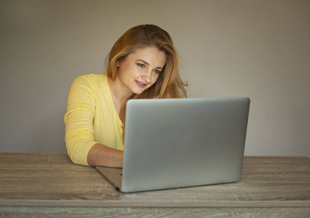 Молодая женщина работает в сети. Девушка с ноутбуком. Улыбающаяся женщина в жёлтом кардигане
 - Фото, изображение