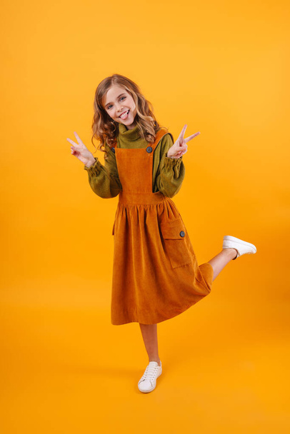 Full length of a smiling little girl wearing a dress standing isolated over blue background, showing peace gesture - Foto, Imagen