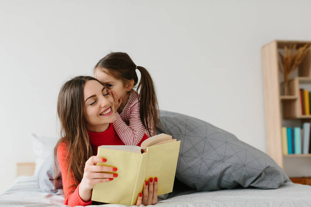 Happy family mother and child daughter reading holding book lying in bed, smiling mom baby sitter telling funny fairy tale to cute preschool kid girl having fun laughing together with bedtime stories - Фото, зображення