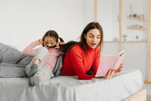 Ευτυχισμένη οικογένεια μητέρα και παιδί κόρη ανάγνωση κρατώντας βιβλίο που βρίσκεται στο κρεβάτι, χαμογελαστή μαμά baby sitter λέει αστείο παραμύθι σε χαριτωμένο κοριτσάκι προσχολικής ηλικίας διασκεδάζουν γελώντας μαζί με παραμύθια πριν τον ύπνο - Φωτογραφία, εικόνα