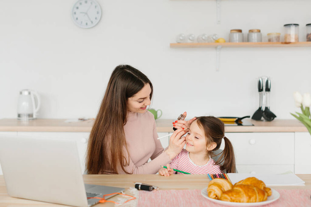 glimlachende mooie vrouw doen make-up terwijl zitten met haar dochtertje in de keuken tijdens het ontbijt. - Foto, afbeelding