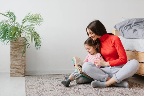 Happy family mother and child daughter reading holding book lying in bed, smiling mom baby sitter telling funny fairy tale to cute preschool kid girl having fun laughing together with bedtime stories - Foto, Bild