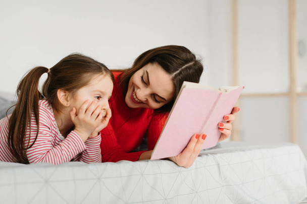 Happy family mother and child daughter reading holding book lying in bed, smiling mom baby sitter telling funny fairy tale to cute preschool kid girl having fun laughing together with bedtime stories - Photo, Image