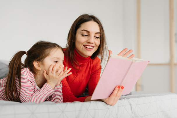 幸せな家族の母親と子供の娘読書本をベッドに寝そべって、笑顔のお母さんの赤ちゃんの妹はかわいい未就学児の女の子に面白いおとぎ話を伝える楽しい時間を過ごす物語と一緒に笑って - 写真・画像