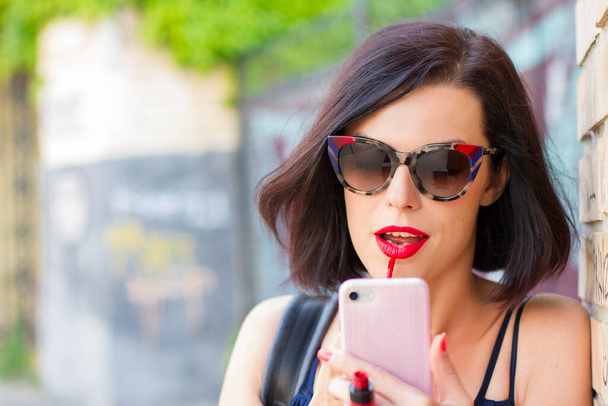 sexy brünette Frau bemalt ihre Lippen mit rotem Lippenstift, Dame benutzt Smartphone als Spiegel. attraktives Mädchen im Freien mit Sonnenbrille. - Foto, Bild