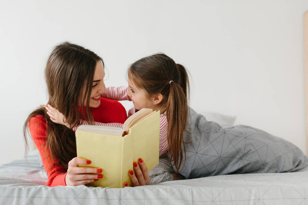 Szczęśliwa rodzina matka i dziecko córka czytanie gospodarstwa książki leżące w łóżku, uśmiechnięta mama opiekunka do dziecka opowiadając zabawną bajkę do cute przedszkola dziewczynka o zabawy śmiejąc się razem z opowieści przed snem - Zdjęcie, obraz