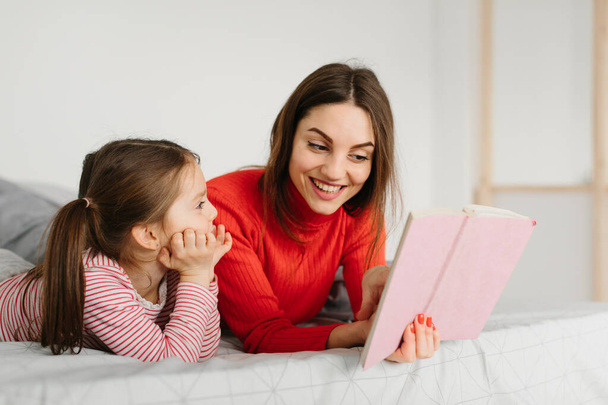 Happy family mother and child daughter reading holding book lying in bed, smiling mom baby sitter telling funny fairy tale to cute preschool kid girl having fun laughing together with bedtime stories - Photo, image