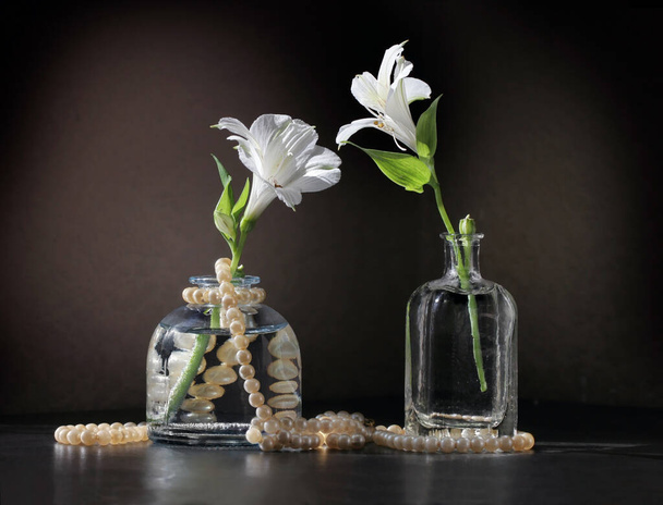 Retro style still life with two Alstroemeria flowers in the small vintage bottles and string of pearls  against a low key background. Selective and soft focus.  - Foto, Bild