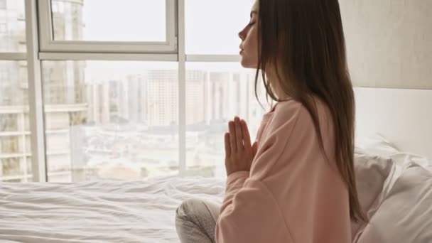 A calm young attractive girl is doing meditation sitting in the bed and holding hands in a praying pose at home - Video