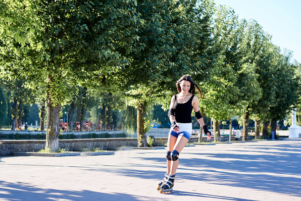 Young pretty brunette woman, riding roller blades in city park in the morning. Fit sporty girl, wearing black top and white shorts, roller skating in action. Full-length portrait of slim sportswoman, - Photo, image