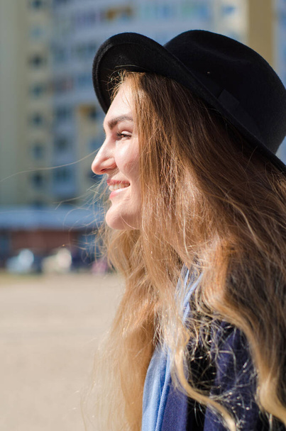 Young caucasian happy smiling girl, in coat and hat in spring city, under rays of spring sun, in background of building - Foto, Imagen