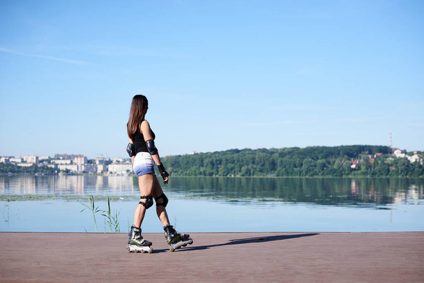 Young pretty brunette woman, with long hair, rollerblading with protective equipment on, on platform by city lake. Full-length portrait of fit slim roller skater girl in summer. - Zdjęcie, obraz