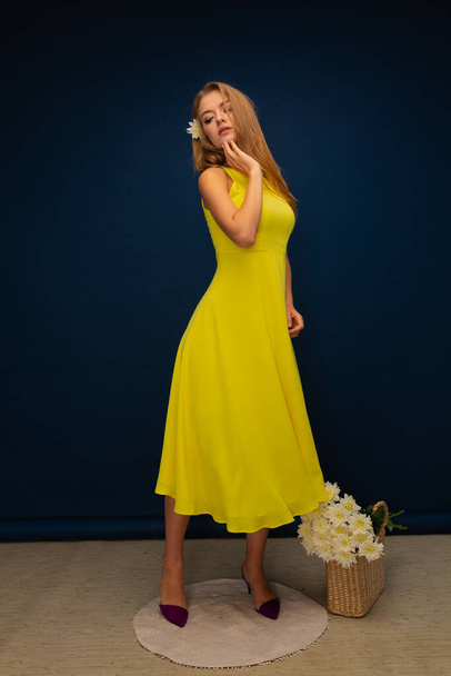 jong mooi blond meisje in gele zomer jurk en gele schoenen met bloemen op blauwe studio achtergrond - Foto, afbeelding