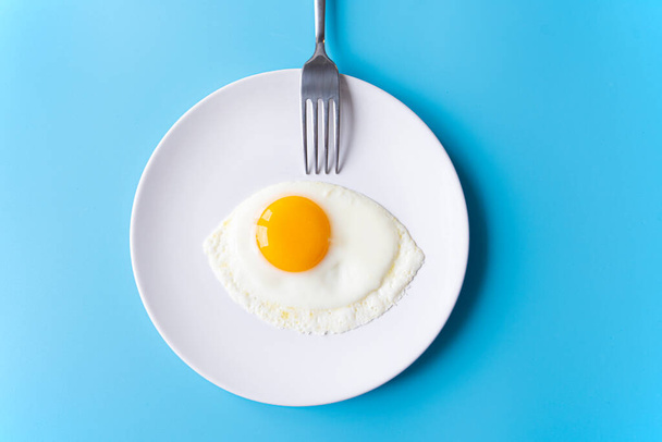 Breakfast, fried egg, yolk, omelet and fork on white plate with color table, creative picture - Foto, Imagen