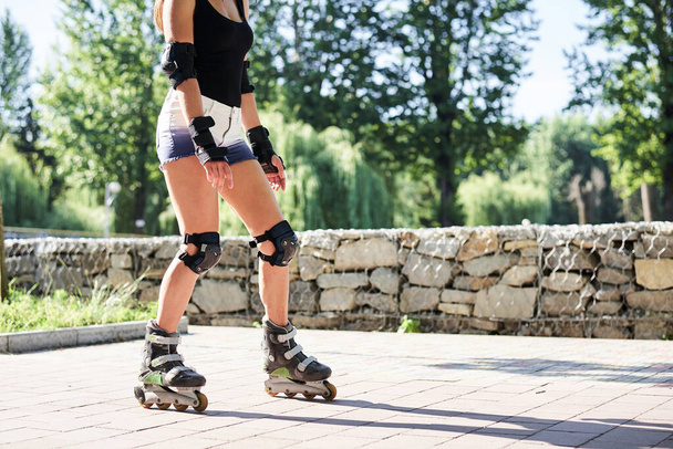 Young woman, roller-blading park with green trees in summer morning at sunny weather. Tan legs, wearing roller-skates and protective equipment, showing the process of riding in town. - Fotoğraf, Görsel